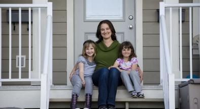 Woman sitting on front porch stairs with two young girls sitting on either side, smiling at the camera
