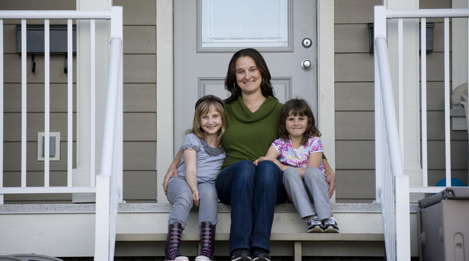 Woman sitting on front porch stairs with two young girls sitting on either side, smiling at the camera